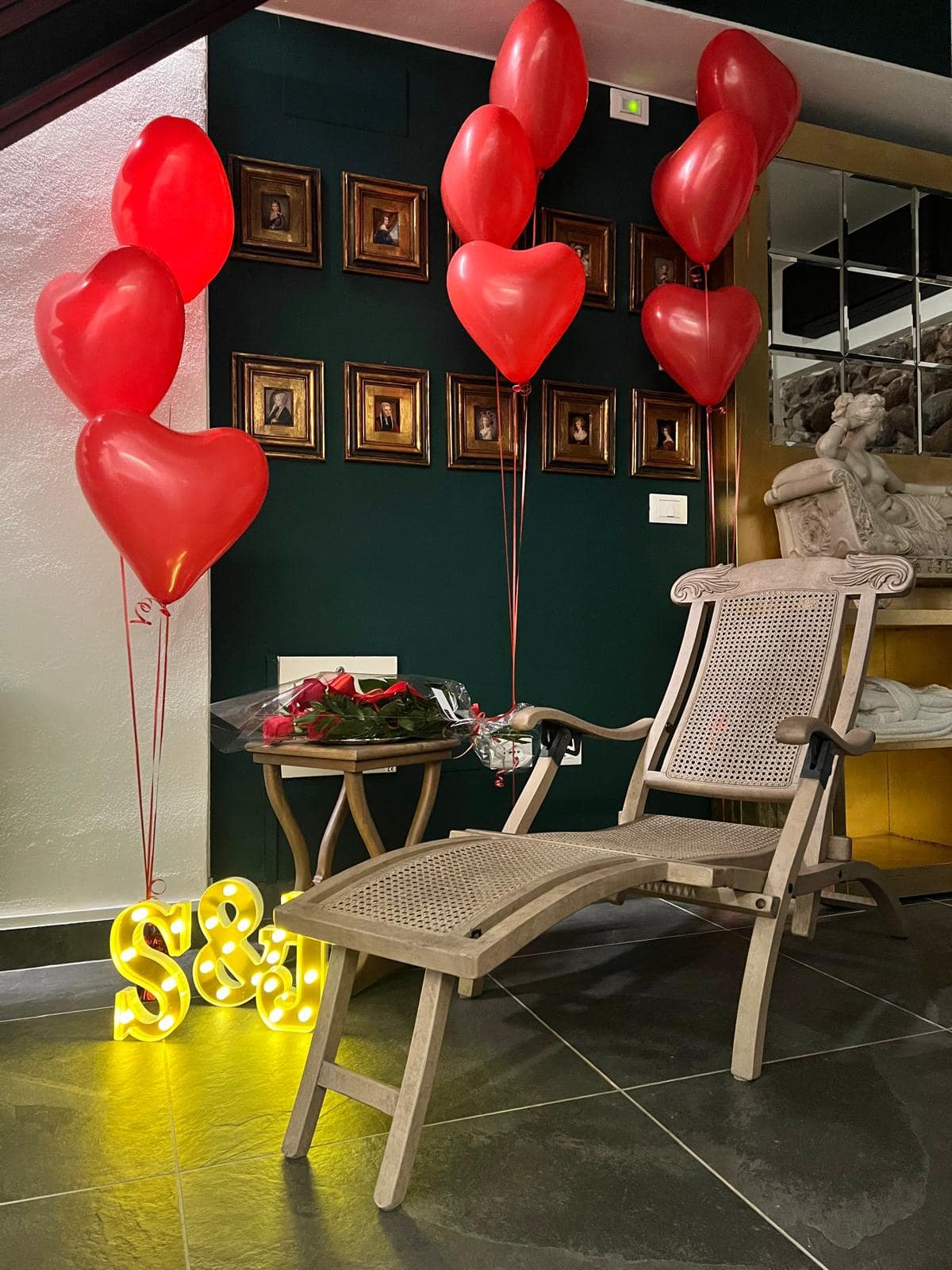 Allestimento a sorpresa al Marcello Sleep & Spa di Verona: palloncini a cuore rossi, bouquet di rose rosse e lettere luminose con le iniziali accanto alla chaise longue d’epoca