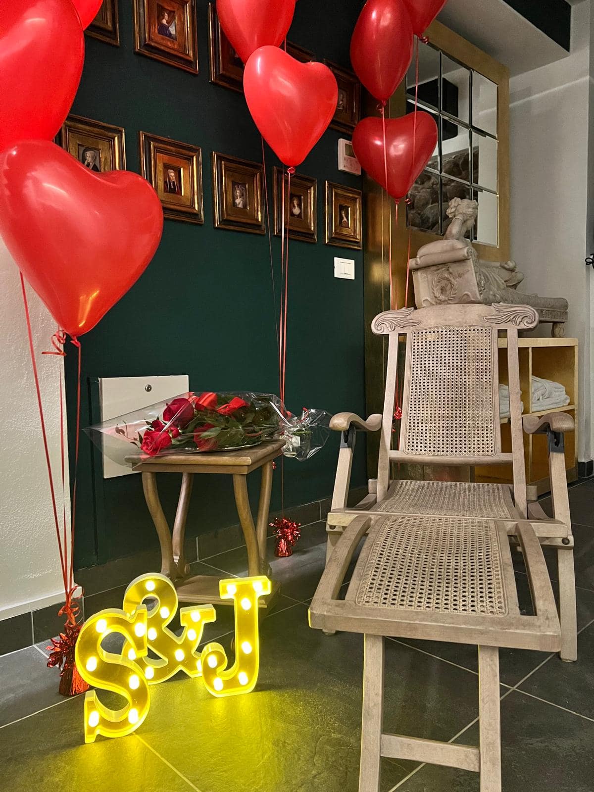 Sorpresa romantica nella suite Marcello a Verona: grappolo di palloncini a cuore rossi, rose rosse sul tavolino e iniziali luminose vicino alla poltrona sdraio in legno
