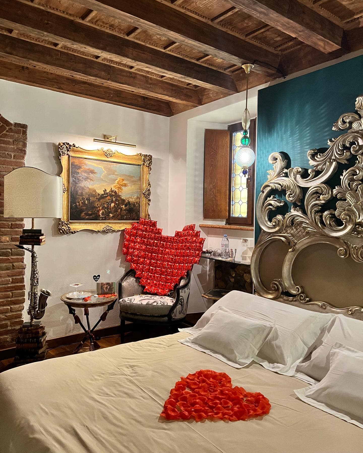 Suite Marcello a Verona allestita per occasione speciale: cuore di petali di rosa sul letto matrimoniale, cuore rosso sulla poltrona, testiera barocca e travi a vista