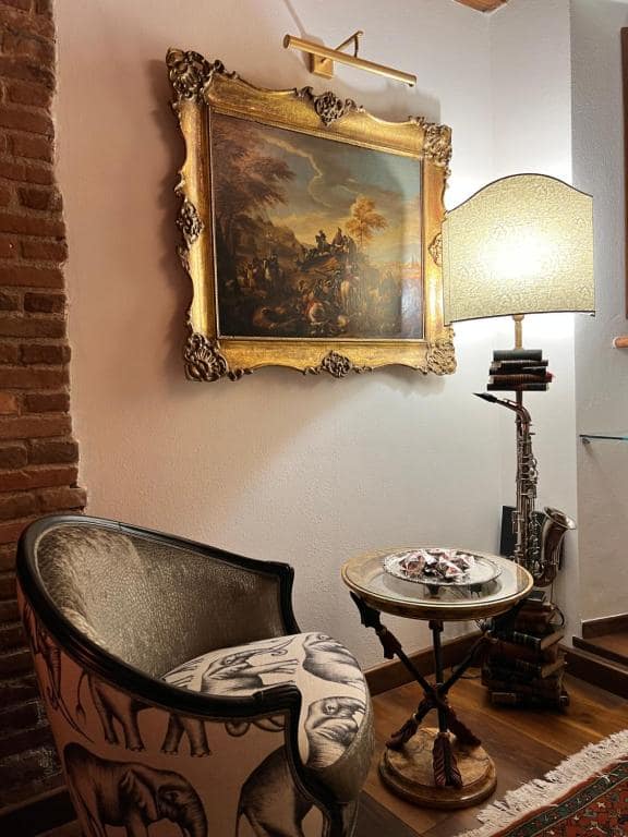 Angolo lettura della suite Marcello a Verona: tela paesaggistica del Settecento in cornice dorata con applique in ottone, lampada su libri d’arte, sassofono d’ottone e poltrona con stampa elefanti Fornasetti-style