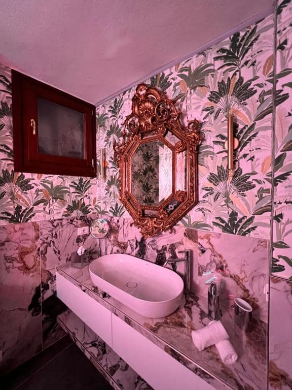 Bagno privato della suite Marcello a Verona in marmo Calacatta viola con venature dorate: lavabo ovale bianco, grande specchio barocco ottagonale dorato, carta da parati tropicale a foglie di banano e applique laterali