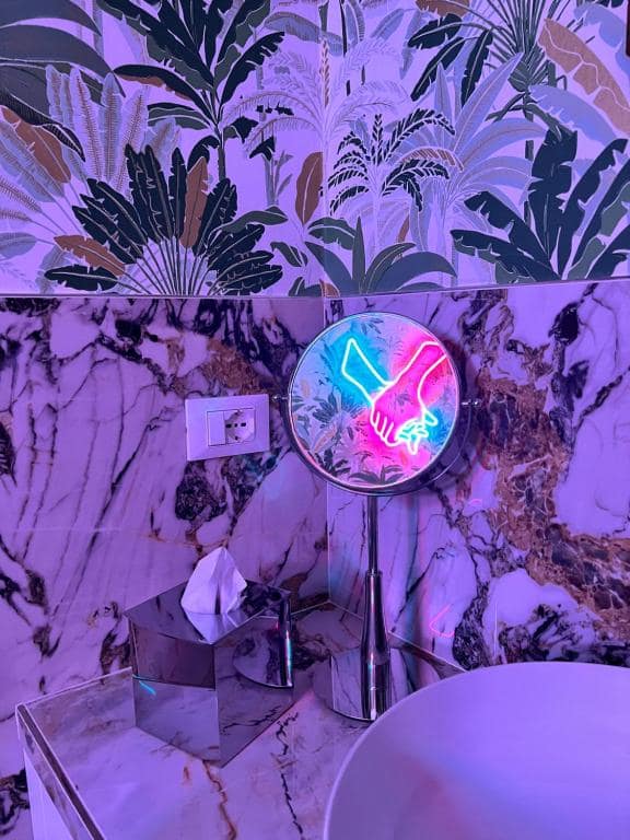 Closeup del bagno privato Marcello a Verona: specchio circolare in metallo che riflette l’insegna al neon a forma di mani intrecciate (rosa e azzurra), dispenser sapone in argento e porta-fazzoletti su piano in marmo Calacatta viola