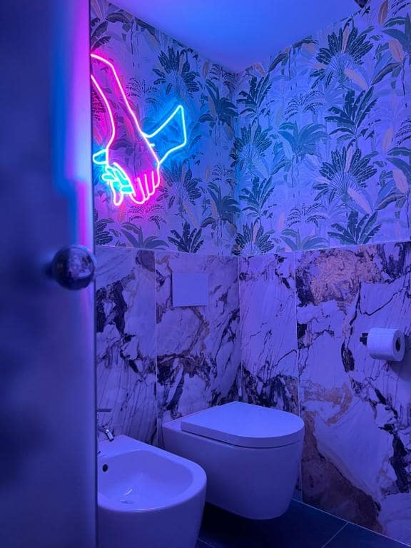 Vista d’insieme del bagno privato Marcello a Verona: WC e bidet, marmo Calacatta viola con venature dorate, insegna al neon a forma di mani intrecciate rosa e azzurra, specchio in legno appeso e carta da parati tropicale