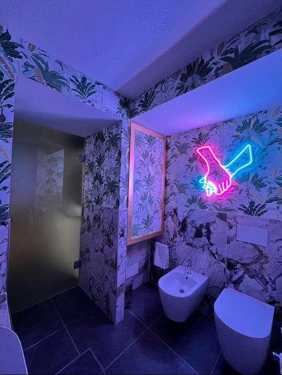 Bagno della suite Marcello a Verona in luce viola: WC e bidet su parete in marmo Calacatta venato d’oro, insegna al neon a forma di mani intrecciate rosa e azzurra e carta da parati tropicale