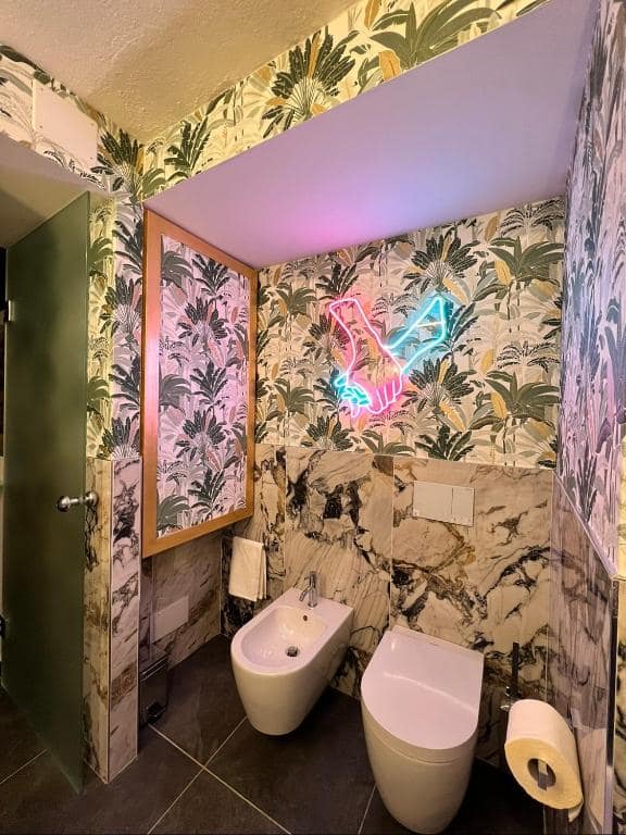 WC e bidet del bagno privato Marcello a Verona: parete in marmo Calacatta viola venato d’oro, insegna al neon a forma di mani intrecciate rosa e azzurra sopra, specchio in legno appeso e carta da parati tropicale