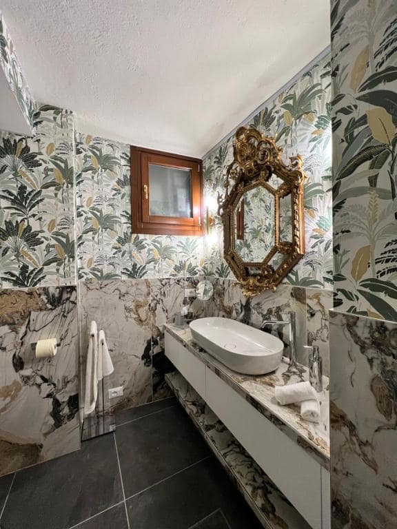 Bagno della suite Marcello a Verona alla luce naturale del giorno: specchio barocco ottagonale dorato, carta da parati a foglie di banano, finestra in legno e lavabo in marmo Calacatta