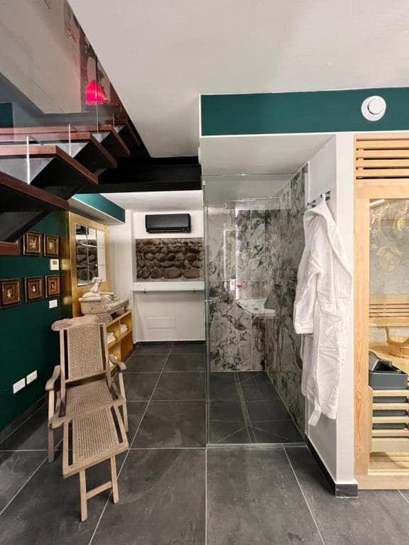 Zona relax della SPA Marcello al piano −1: scala in legno sopra, parete verde smeraldo con galleria di miniature, doccia in marmo, accappatoi bianchi appesi e sedia in vimini in primo piano