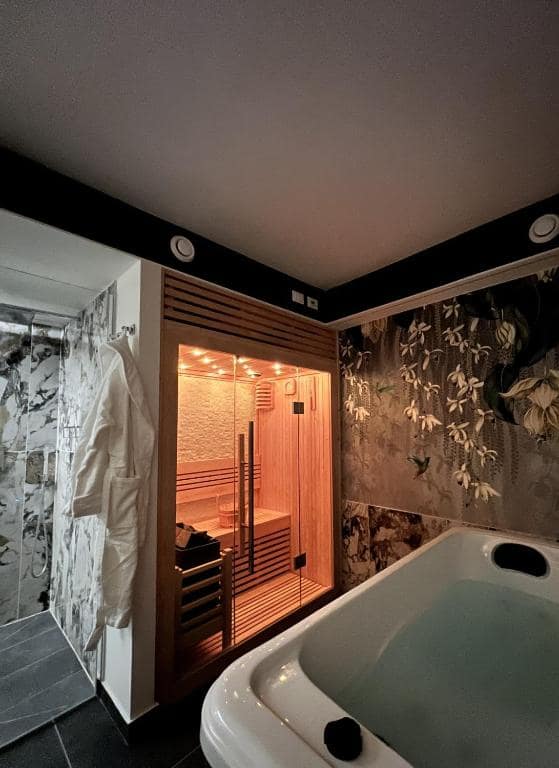 Sauna finlandese in luce calda della SPA privata Marcello a Verona, accanto alla vasca idromassaggio: doccia in marmo, accappatoio bianco appeso e carta da parati botanica scura