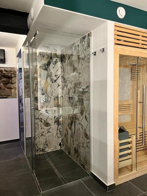 Doccia con box in vetro della SPA privata Marcello a Verona: pareti in marmo venato grigio-verde con venature dorate, soffione a pioggia e sauna finlandese in legno chiaro a destra