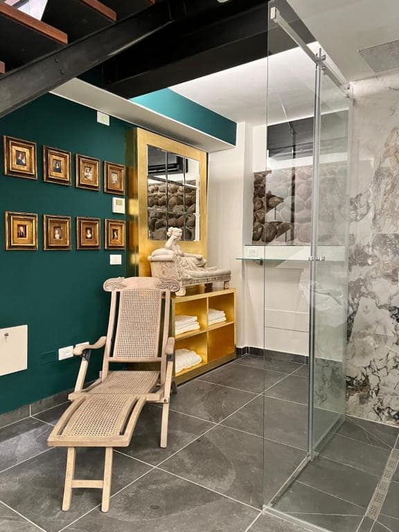 Zona relax e doccia in marmo della SPA Marcello a Verona: parete verde smeraldo con galleria di miniature in cornice dorata, statua di Venere su console dorata e sedia in vimini in primo piano