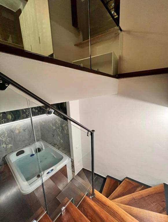 Scala in legno con balaustra in vetro che dalla suite Marcello al piano terra scende alla SPA privata al piano −1, vista dall’alto, con la vasca idromassaggio in basso e carta da parati botanica