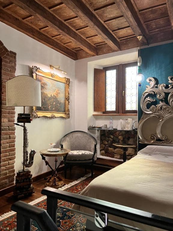 Vista della suite Marcello a Verona dal letto: arco originale in mattoni del Settecento, tela paesaggistica in cornice dorata, finestra in legno con vetri colorati e poltrona con sassofono