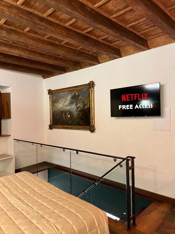 Soppalco della suite Marcello a Verona: tela paesaggistica del Settecento in cornice dorata accanto alla Smart TV con accesso Netflix gratuito, balaustra in vetro e soffitto a travi a vista