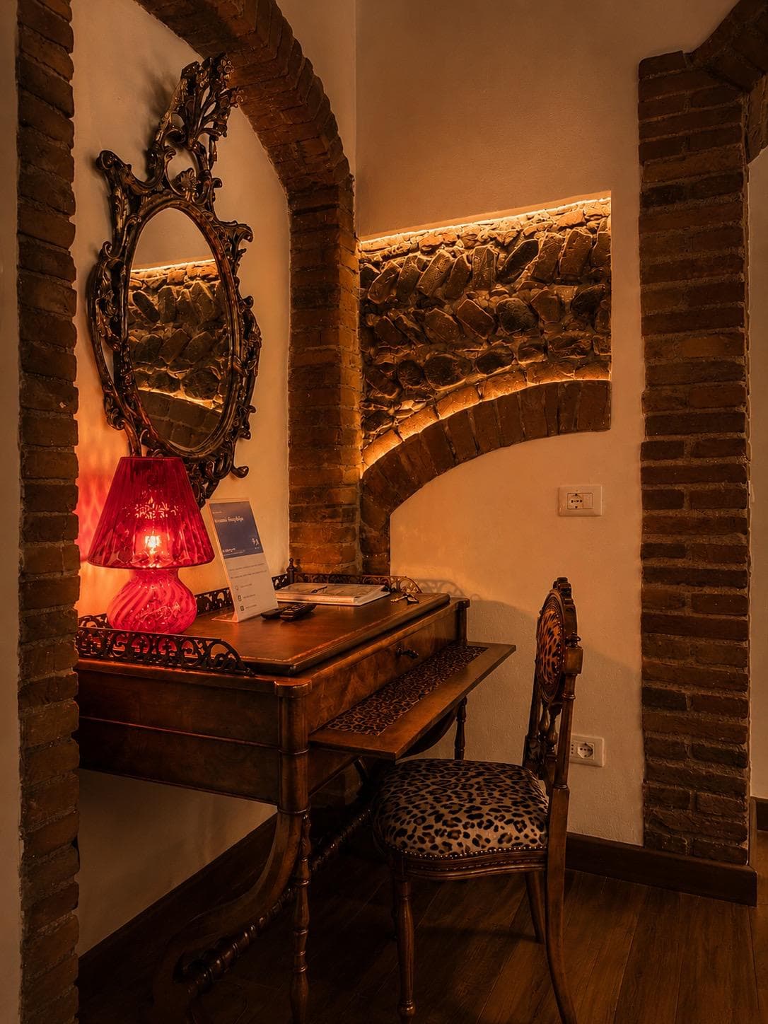 Arco originale in mattoni del Settecento nella suite Marcello a Verona: scrittoio antico in legno noce, specchio barocco ovale dorato, lampada da tavolo in vetro rosso e sedia con stampa leopardata