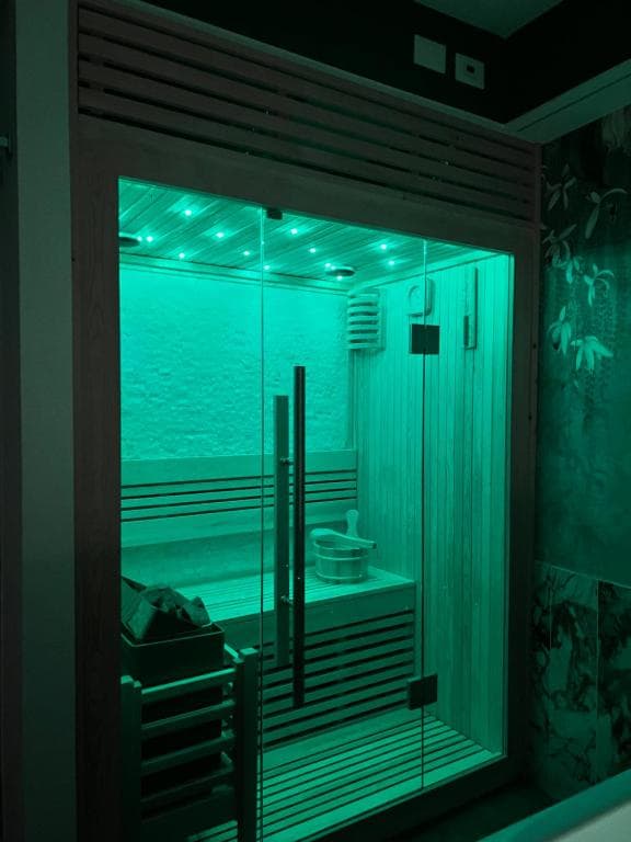 Sauna finlandese privata della SPA Marcello a Verona: porta in vetro con cornice in legno chiaro, interno illuminato da cromoterapia LED in verde brillante, panche a due livelli e secchiello in legno