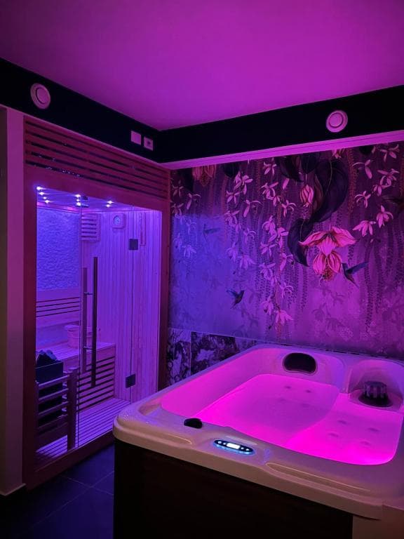 SPA privata Marcello a Verona in atmosfera Aurora: vasca idromassaggio in cromoterapia rosa intensa, sauna finlandese sullo sfondo e carta da parati botanica saturata di rosa