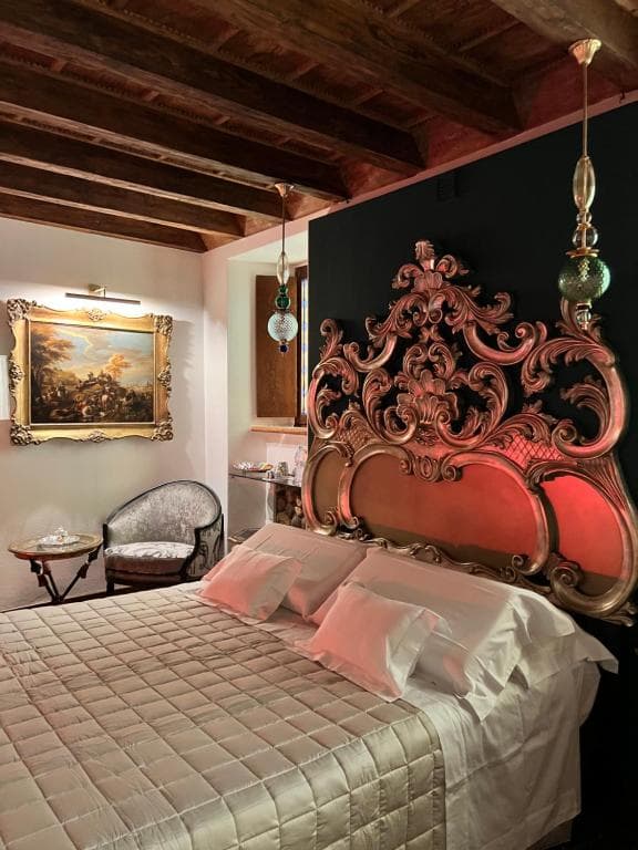 Suite Marcello a Verona di sera: letto matrimoniale con testiera barocca dorata, tela paesaggistica del Settecento in cornice dorata illuminata, lampade pendenti in vetro di Murano e soffitto a travi a vista