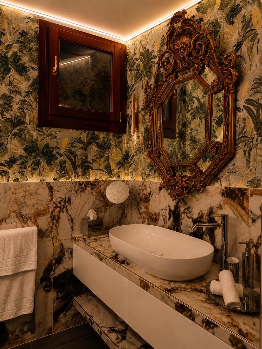Baño privado de la suite Marcello en Verona en mármol Calacatta violeta con vetas doradas: lavabo ovalado blanco, gran espejo barroco octogonal dorado, papel pintado tropical de hojas de banano y apliques laterales