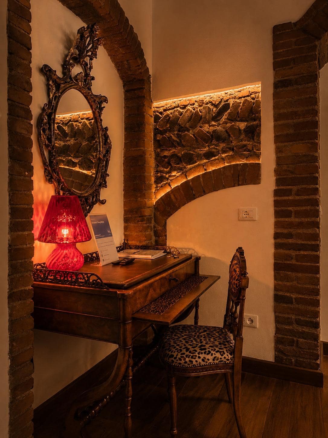 Arco original en ladrillo del siglo XVIII en la suite Marcello en Verona: escritorio antiguo de nogal, espejo barroco oval dorado, lámpara de mesa de cristal rojo y silla con estampado de leopardo